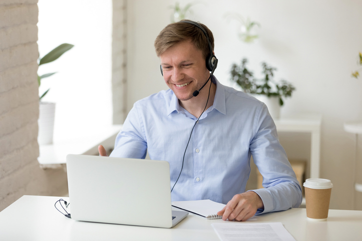 Smiling man in headphones looking at computer screen watching webinar, making business video call, young businessman consulting remote client online writing notes, hr holding distance job interview