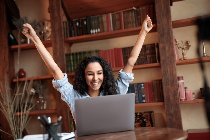 Great News Concept. Portrait of emotional ecstatic woman celebrating online win sitting at desk, using laptop computer, copy space. Smiling happy female model got promotion, raising hands up