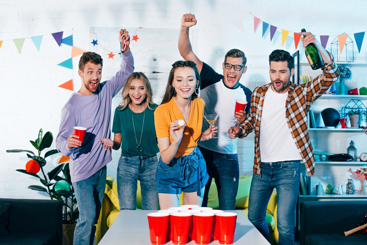 happy young friends looking at smiling girl playing beer pong at home party
