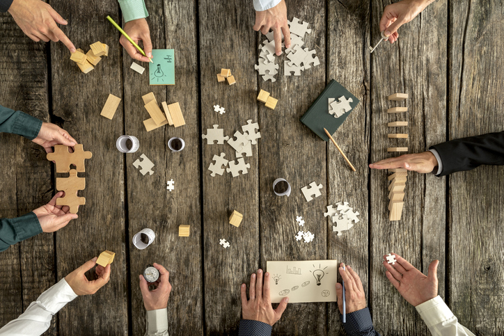 Business planning and brainstorming concept with a team of ten businessmen organizing strategy while holding puzzle pieces, writing down ideas on paper and rearranging wooden blocks, top view.