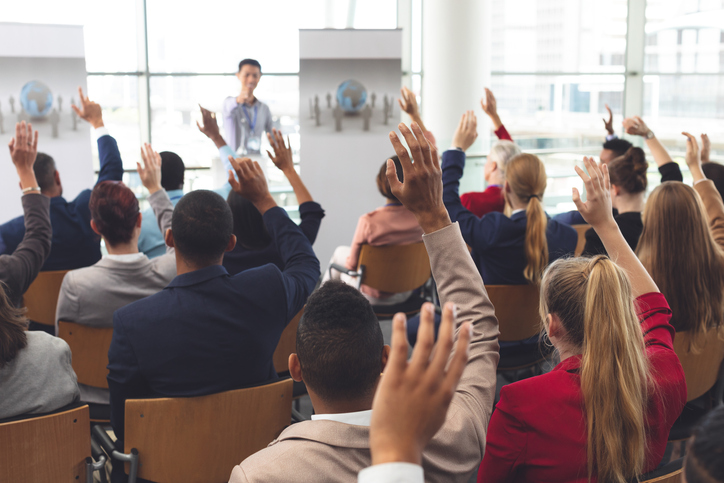 Rear view of diverse business people raising hands while they are sitting in front of Asian businessman at business seminar in office building