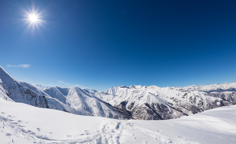 Sun star glowing over snowcapped mountain range and high mountain peaks in the italian alpine arc, in a bright sunny day of winter. Candid snowy slope in the foreground.
